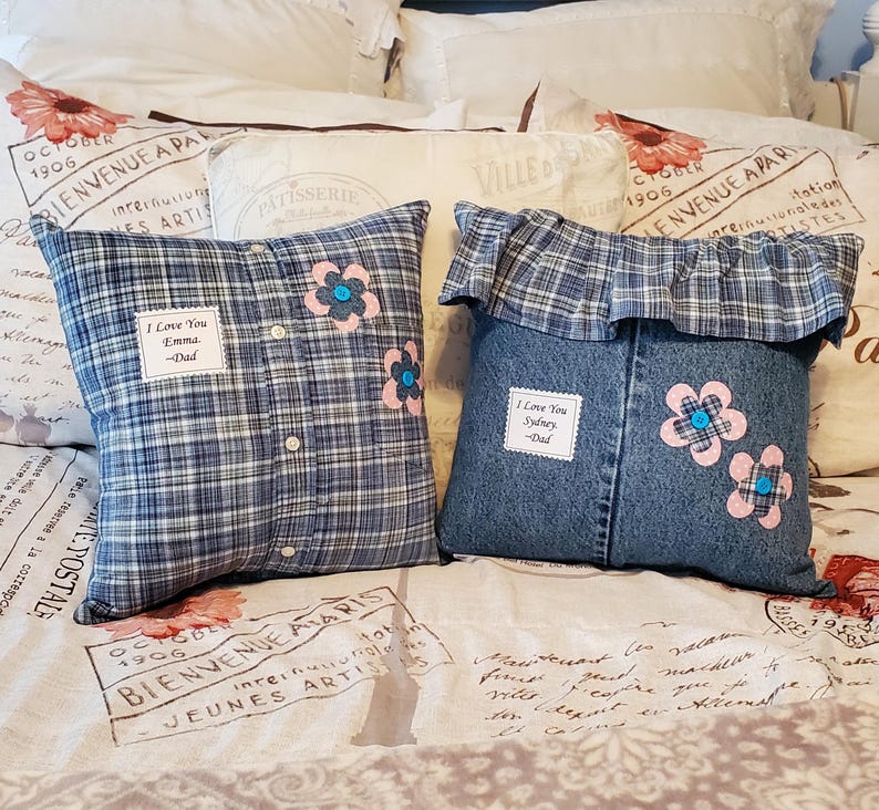 Two decorative pillows made from clothing. One is made from a blue plaid shirt, the other from denim. Both have appliqued pink flowers with blue centers. The plaid pillow has the text "I Love You Emma - Dad". The denim pillow has the text "I Love You Sydney - Dad".