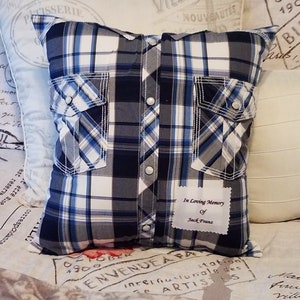 A square pillow made from a blue, white, and gray plaid shirt. The pillow has two pockets and silver snap closures. A white label with the text "In Loving Memory of Jack Franz" is sewn on the front.