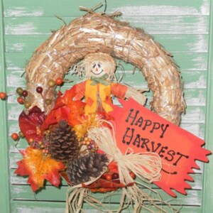 May include: A harvest wreath featuring a straw base, a scarecrow, autumn leaves, pine cones, and berries. A red sign with "Happy Harvest" text is attached. The wreath is set against a green shutter background.