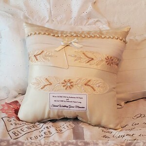 Ivory-colored decorative memory pillow made from wedding dress with embroidered floral and beaded accents. Features a bow and a label with text.