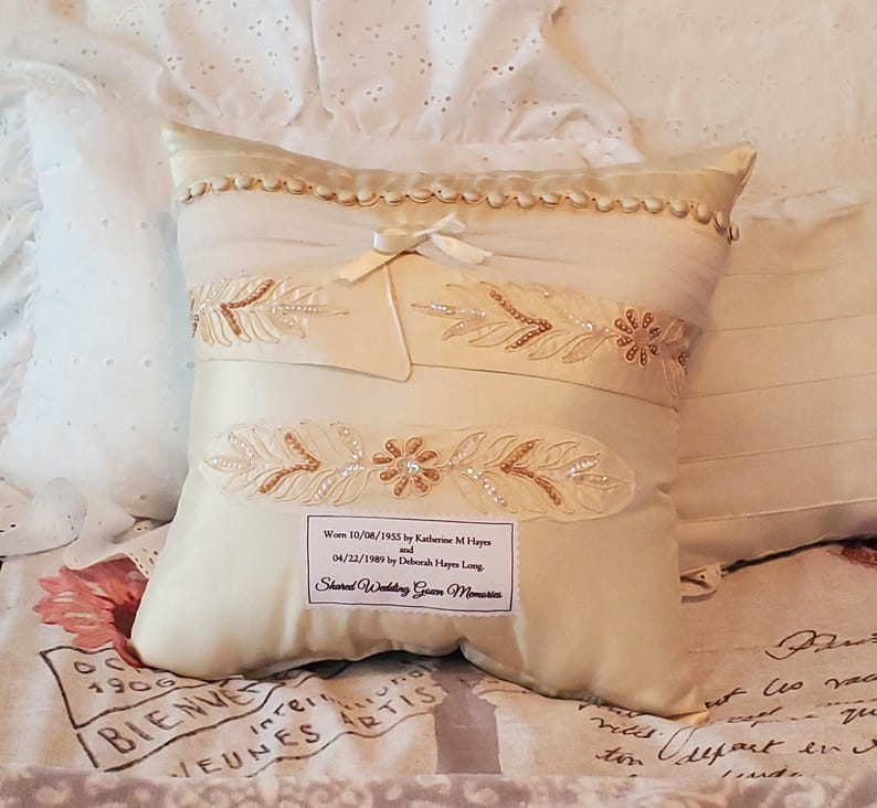 Ivory-colored decorative memory pillow made from wedding dress with embroidered floral and beaded accents. Features a bow and a label with text.