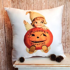 May include: White square pillow featuring a vintage Halloween illustration. The image shows a smiling child in a hat holding a carved orange pumpkin with the words "Halloween Greeting". Decorative item for seasonal home decor.