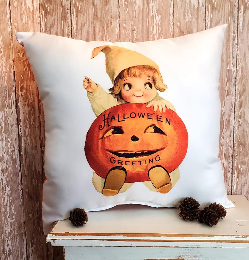 May include: White square pillow featuring a vintage Halloween illustration. The image shows a smiling child in a hat holding a carved orange pumpkin with the words "Halloween Greeting". Decorative item for seasonal home decor.