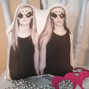 May include: A white pillow with a photo of two girls wearing black tank tops and sunglasses. The girls have long blonde hair and are looking at the camera. The pillow is sitting on a table with a pink sparkly headband.