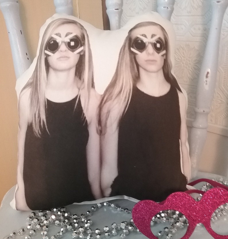 May include: A white pillow with a photo of two girls wearing black tank tops and sunglasses. The girls have long blonde hair and are looking at the camera. The pillow is sitting on a table with a pink sparkly headband.
