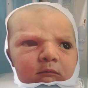 May include: A white pillow with a printed image of a baby's face. The baby has a serious expression and is looking directly at the camera.