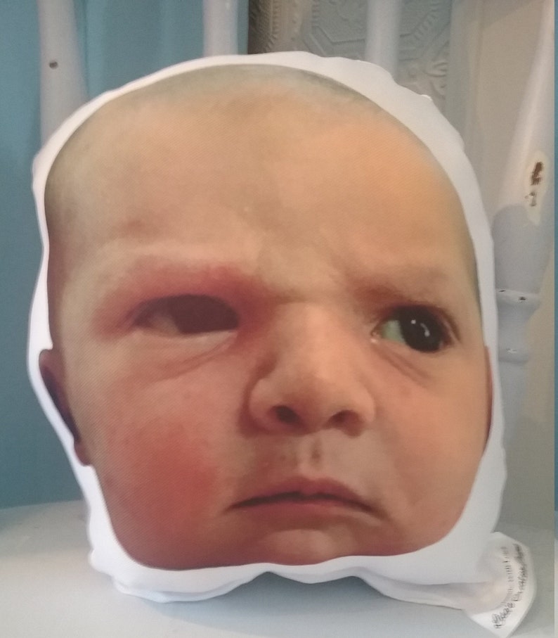 May include: A white pillow with a printed image of a baby's face. The baby has a serious expression and is looking directly at the camera.