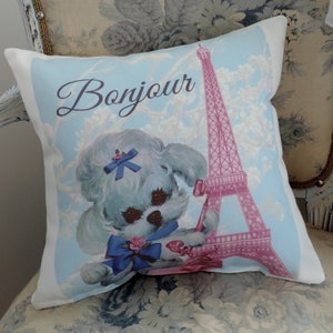 May include: A white pillow with a blue and pink design featuring a cartoon poodle wearing a blue bow and the Eiffel Tower. The text "Bonjour" is written in a cursive font.