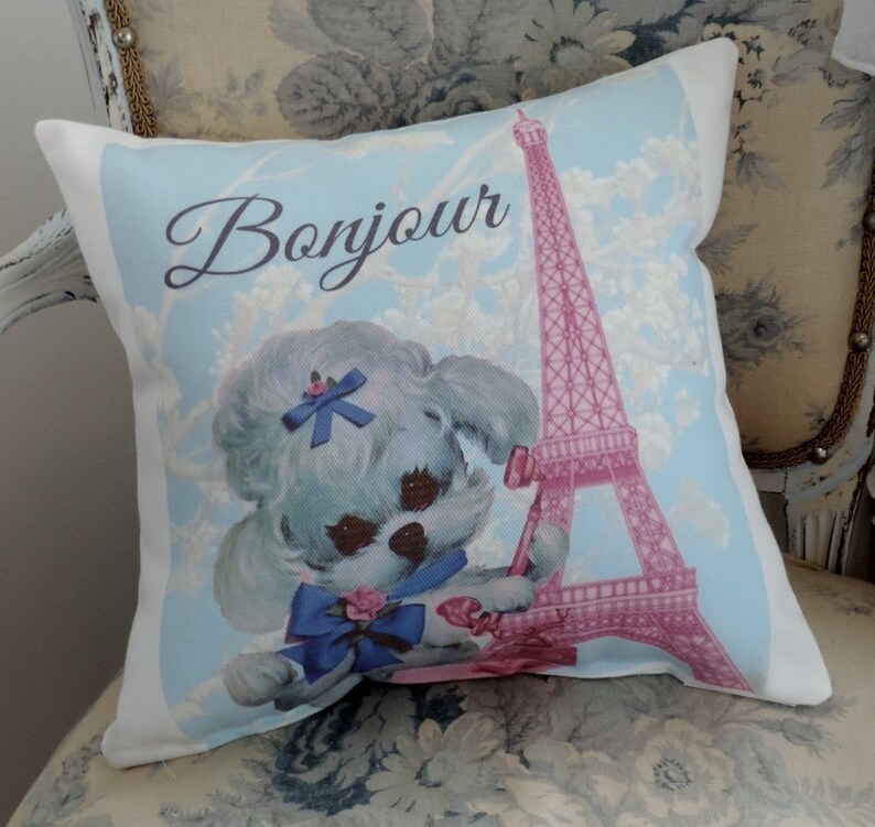 May include: A white pillow with a blue and pink design featuring a cartoon poodle wearing a blue bow and the Eiffel Tower. The text "Bonjour" is written in a cursive font.