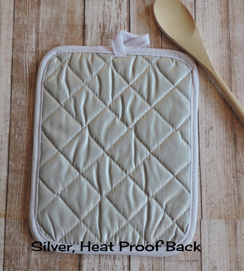 May include: Silver heat-proof pot holder with a quilted design. The back of the pot holder is visible in the image. "Silver, Heat Proof Back" is printed on the bottom edge of the pot holder.