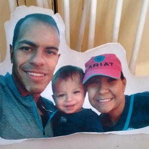 May include: A white pillow with a photo of a family of three. The father is on the left, the mother is on the right, and their child is in the middle. The mother is wearing a pink hat with the word "ARIAT" on it.