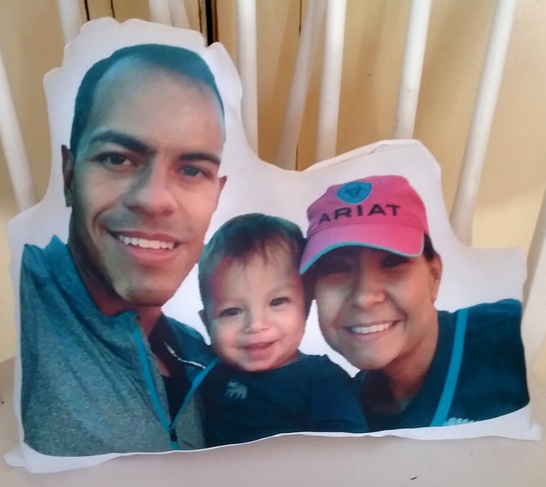May include: A white pillow with a photo of a family of three. The father is on the left, the mother is on the right, and their child is in the middle. The mother is wearing a pink hat with the word "ARIAT" on it.