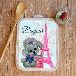 May include: A white pot holder with a blue background featuring a pink Eiffel Tower, a white poodle with a blue bow, and the text "Bonjour".