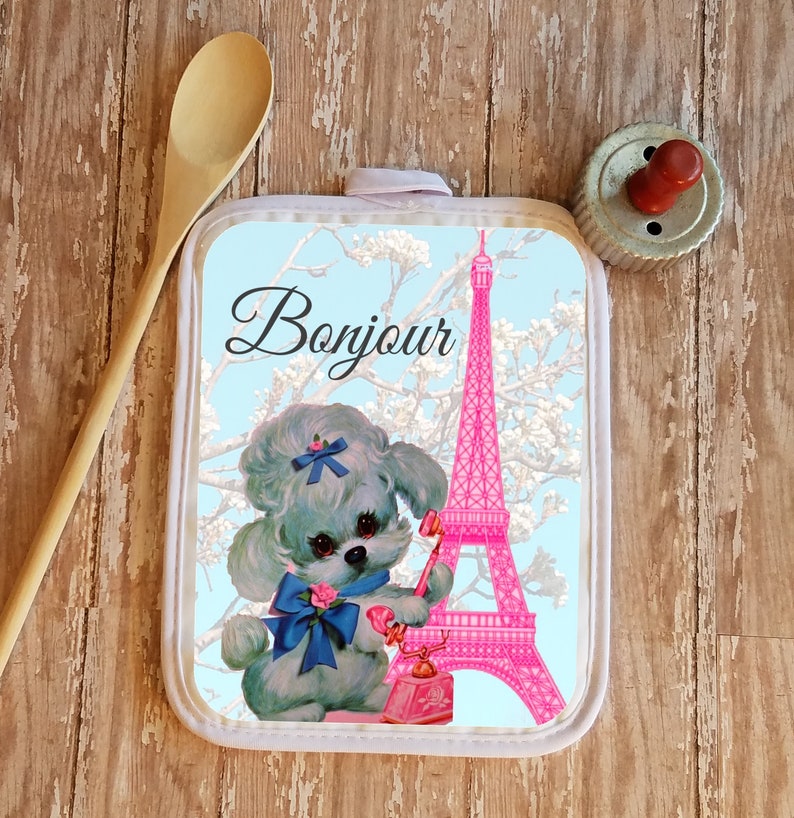 May include: A white pot holder with a blue background featuring a pink Eiffel Tower, a white poodle with a blue bow, and the text "Bonjour".