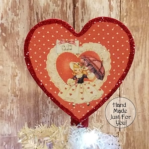 May include: Red glitter heart-shaped ornament with a vintage Valentine's Day image of a girl holding an umbrella. The ornament has white polka dots and a red glitter trim. The text "My Valentine" is visible on the ornament. The text "Hand Made Just For You!" is visible in a circle below the ornament.