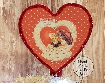 May include: Red glitter heart-shaped ornament with a vintage Valentine's Day image of a girl holding an umbrella. The ornament has white polka dots and a red glitter trim. The text "My Valentine" is visible on the ornament. The text "Hand Made Just For You!" is visible in a circle below the ornament.