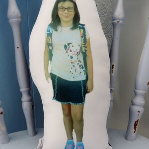 May include: A white pillow with a photo of a young girl wearing a white shirt with a black and white dog graphic, a black skirt, and blue shoes. The girl is wearing a pink flower in her hair and has glasses on.