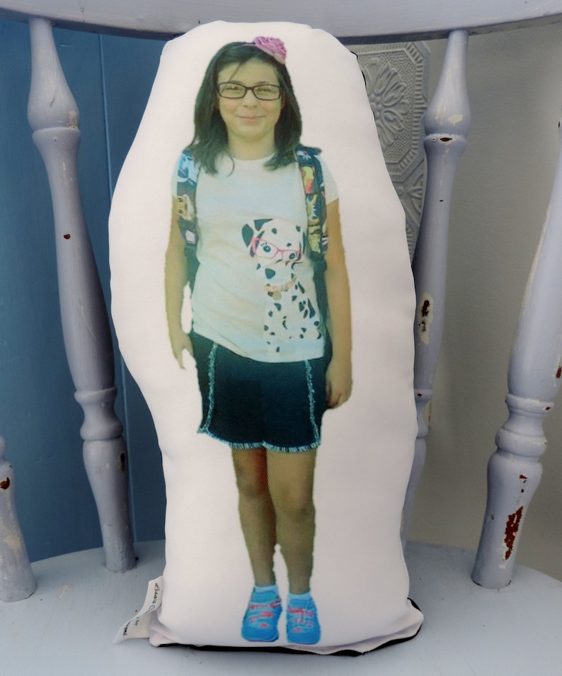 May include: A white pillow with a photo of a young girl wearing a white shirt with a black and white dog graphic, a black skirt, and blue shoes. The girl is wearing a pink flower in her hair and has glasses on.