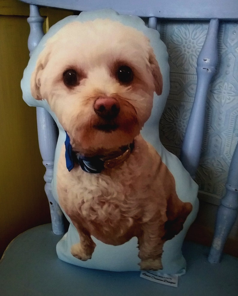 Pu&ograve; includere: Un cuscino a forma di cane bianco e marrone con un papillon blu. Il cuscino &egrave; seduto su una sedia blu.