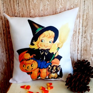 May include: White square Halloween throw pillow featuring a vintage illustration of a smiling witch holding a jack-o'-lantern and broom, with a raccoon. The witch wears a black hat and cape, with a blue skirt. Candy corn is scattered in the foreground.