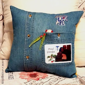 Blue denim pillow made from a shirt with a pocket and button details. The pillow has a UK logo patch and a photo with the text "Proud Grandpa." Two green and red fishing lures are in the pocket.