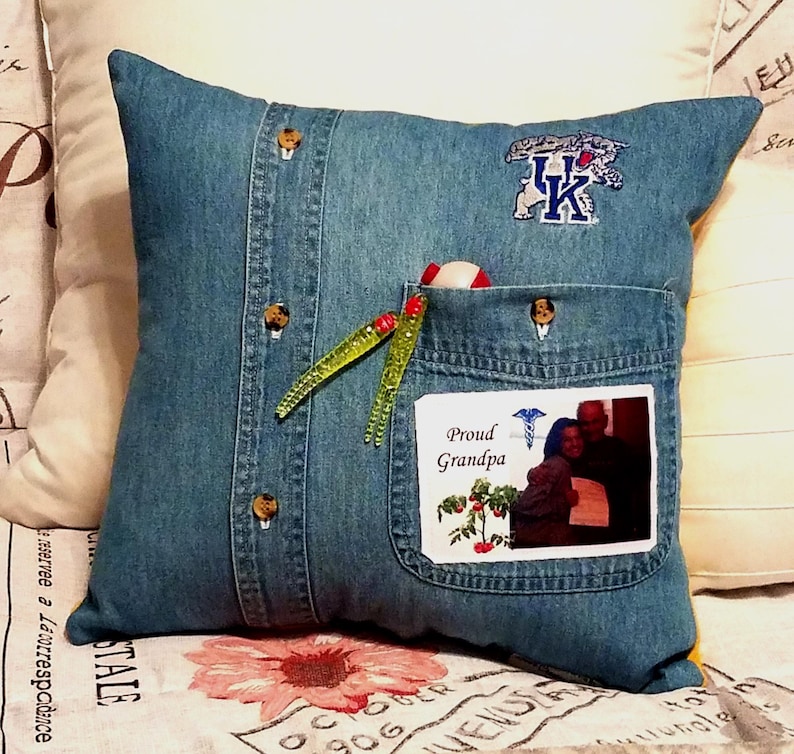 Blue denim pillow made from a shirt with a pocket and button details. The pillow has a UK logo patch and a photo with the text "Proud Grandpa." Two green and red fishing lures are in the pocket.
