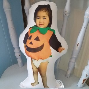 May include: A white pillow with a photo of a child dressed as a pumpkin for Halloween. The child is wearing a green and orange pumpkin costume and a white diaper. The pillow is sitting on a white chair.