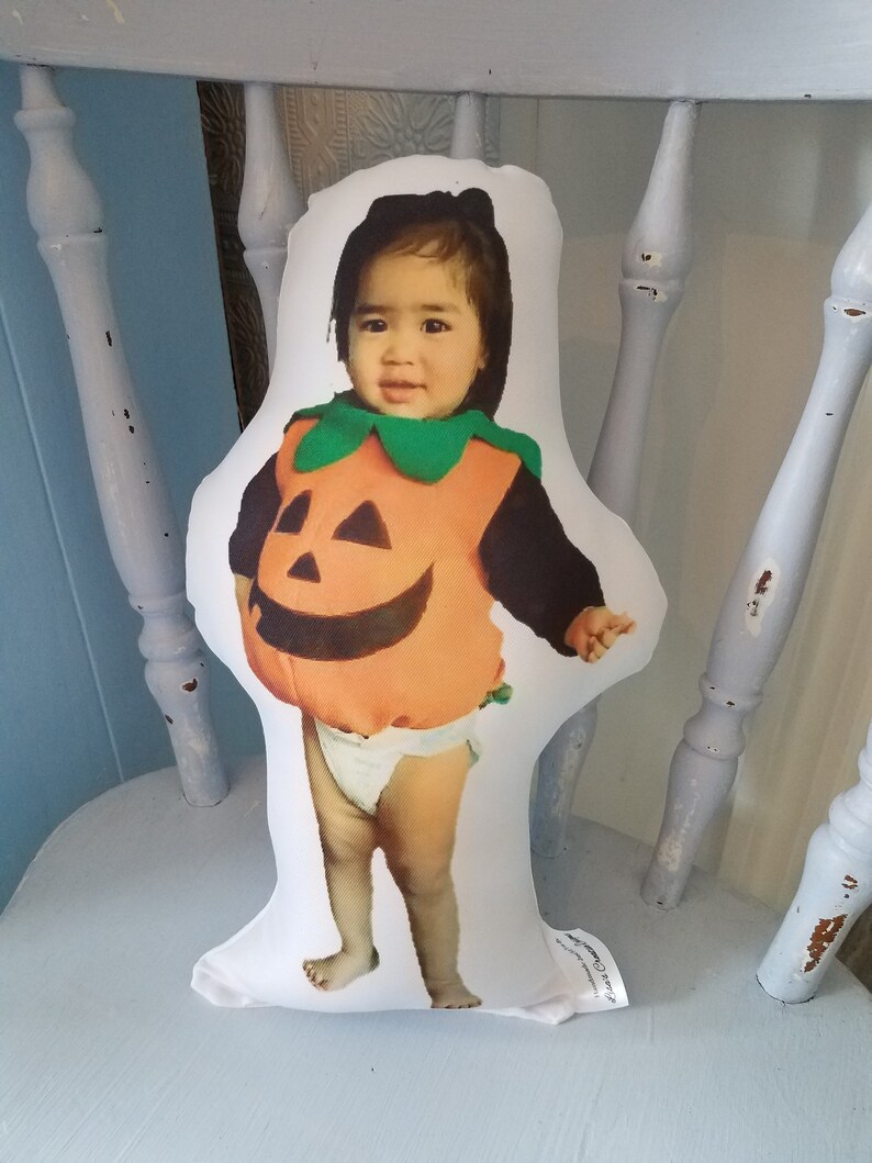 May include: A white pillow with a photo of a child dressed as a pumpkin for Halloween. The child is wearing a green and orange pumpkin costume and a white diaper. The pillow is sitting on a white chair.