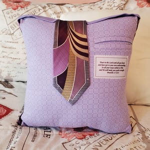 A lavender pillow made from a shirt with a patterned design, featuring a dark purple and gold tie. The pillow has a pocket detail and a label with text from Proverbs 3:5-6.