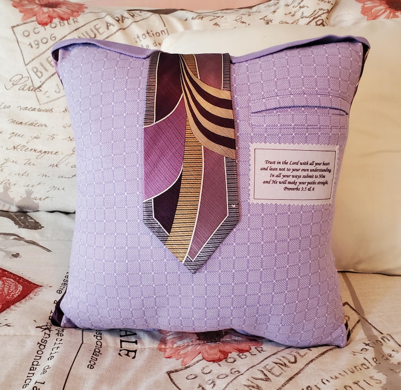 A lavender pillow made from a shirt with a patterned design, featuring a dark purple and gold tie. The pillow has a pocket detail and a label with text from Proverbs 3:5-6.
