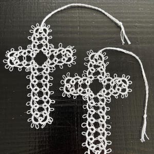 May include: Two white cross-shaped ornaments crafted from intricate tatting lace. Each cross features a repeating looped pattern and includes a thin white thread loop at the top for hanging. The delicate, openwork design creates a detailed texture.