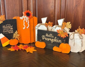 May include: A collection of wooden fall-themed decorations, including orange pumpkins, decorative leaves, and signs. One sign reads "Thankful Blessed Pumpkin Bliss," and another says "Hello Pumpkin." The pumpkins vary in size and style.