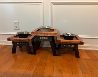 May include: Three wooden pet food stands with stainless steel bowls. The stands are stained in a dark brown and black color scheme. Each stand has a name engraved on it: Evie, Remmi, and Charlie.