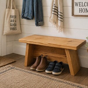 May include: A wooden bench with a natural finish sits in a hallway. Two pairs of shoes are placed underneath the bench. A tote bag with the text "FRESH AIR & GOOD VIBES" hangs on the wall. A sign that says "WELCOME HOME" is also on the wall.