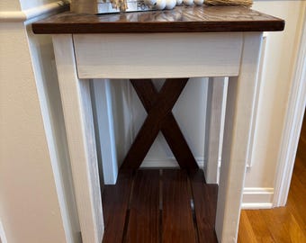 May include: White end table with a brown top and a lower shelf. The table has an X-shaped support in the middle. The table is decorated with a stack of books, a candle, and a string of white beads.