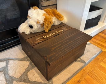 May include: A dark stained wooden toy box with the text "Remi's Toys" engraved on the lid. A plush dog toy with white and brown fur rests on top. The box is rectangular and sits on a stone surface.