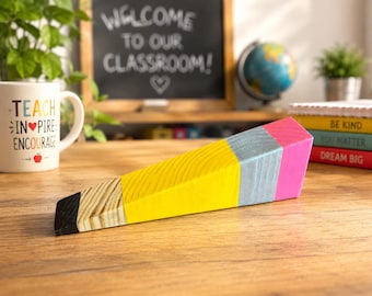 May include: A wooden doorstop shaped like a large pencil, featuring a black tip, a natural wood grain section, a bright yellow body, a light blue band, and a pink eraser end. The object is positioned horizontally on a wooden surface.