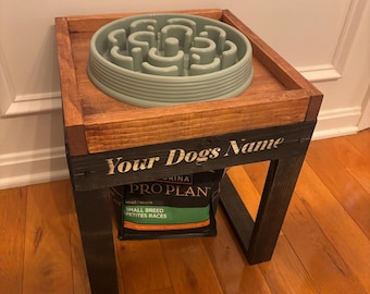 May include: A wooden dog bowl stand with a light green slow feeder bowl on top. The stand has a dark stained frame and the text "Your Dogs Name" is printed on the front. A bag of dog food is visible below.