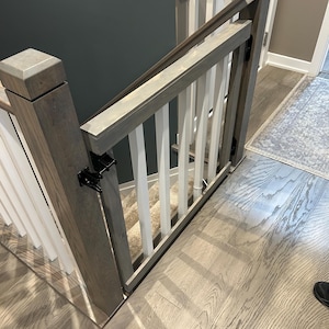 May include: A wooden baby gate installed at the top of a staircase. The gate is made of light gray wood with white vertical spindles. The gate is closed and secured with black hinges. The staircase has light-colored wooden steps and a beige carpet runner.