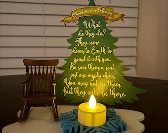May include: A Christmas-themed decorative piece featuring a green tree with the text "Christmas in Heaven." A small rocking chair and a candle in a blue snowflake holder sit on a white base. The tree has a gold star on top.