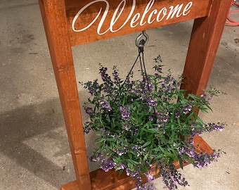 May include: A wooden welcome sign with a hanging planter. The sign is stained a dark brown color and has the word "Welcome" written in white cursive lettering. The planter is hanging from a metal hook and contains a green plant with purple flowers.