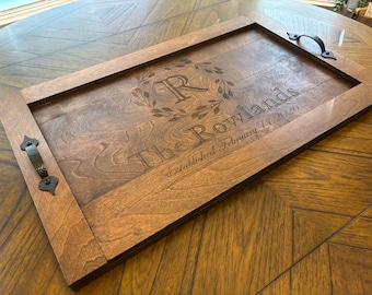 May include: A rectangular wooden serving tray with a dark stain. The tray features a decorative border and handles. The center is engraved with a family name, initial, and date. The handles are dark metal.