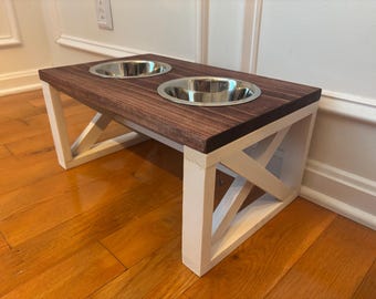 May include: A raised dog feeder with two stainless steel bowls set into a dark stained wooden top. The base is white with an X-shaped design on the sides. The feeder is designed to elevate the bowls for easier access.