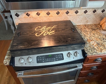 May include: A dark stained wooden stove top cover with the words "Chef Joe" in a cursive font. The cover has two small black handles and is resting on a stainless steel stove.