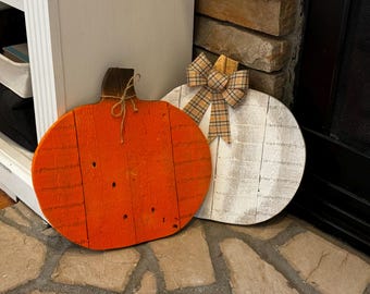 May include: Two wooden pumpkins, one painted orange and one painted white. The white pumpkin has a plaid bow on top.