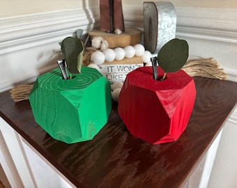 May include: Two wooden apple-shaped decorations, one green and one red, sit side-by-side. Each has a dark green leaf and a metal stem. The apples have a faceted design and are placed on a dark brown surface.
