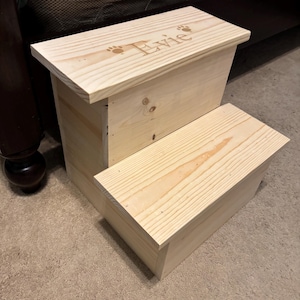 May include: A two-step wooden pet step stool. The natural wood-colored stool has the name "Evie" and paw prints engraved on the top step. The step stool is designed to help pets reach higher places.
