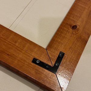 May include: Close-up of a corner joint of a wooden frame with black metal corner brackets. The wood is stained a dark brown colour.