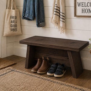 May include: A dark brown wooden bench with angled legs, holding two pairs of shoes. A woven rug and a tote bag with the text "FRESH AIR & GOOD VIBES" are in the foreground. A sign that says "WELCOME HOME" is on the wall.