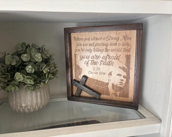 May include: Wooden framed plaque with engraved text: "When you silence a Strong Man...you are afraid of the truth. RIP Charlie Kirk." A small wooden cross rests in front of the plaque. A decorative plant in a beige pot is to the left.